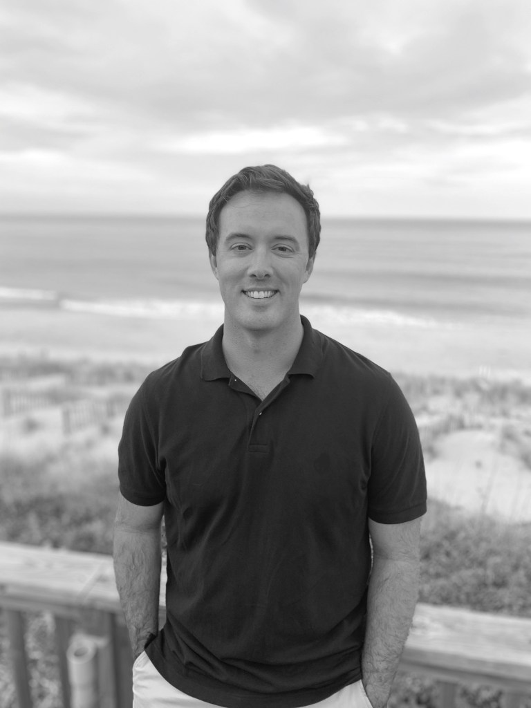 A photo of the author Perrin D Hayes, in black and white in front of a beach and ocean in the background